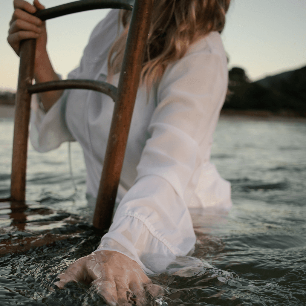 A close-up outdoor scene at the edge of calm water shows a person wading chest-deep or lower near a metal ladder, with one hand gripping a rusted vertical rail and the other hand extended into the water in the foreground.