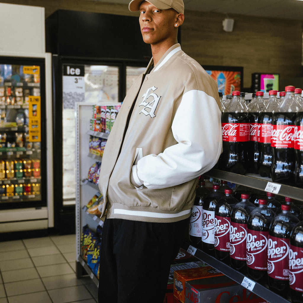 Inside a convenience store or gas-station market, a young man stands in the foreground beside a beverage aisle, framed from mid-thigh up at a slight low angle.