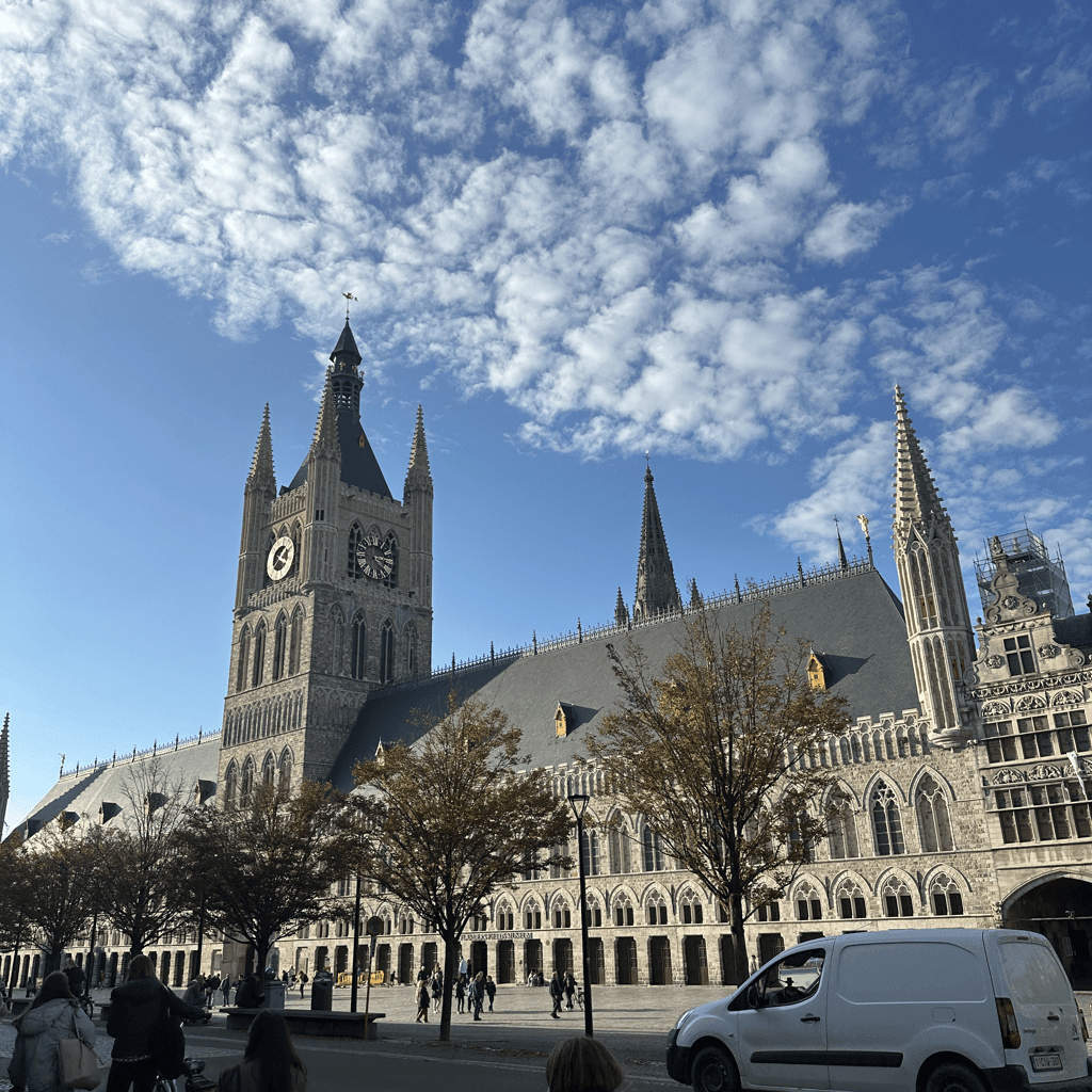 A grand Gothic-style stone building dominates the midground, its vast steep gray slate roof punctuated by dormer windows and topped by a tall central belfry tower with a clock face and pointed corner turrets, alongside slender ornate spires rising toward the right.