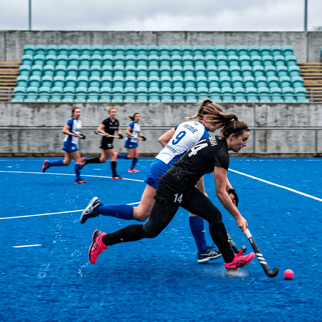 Two field hockey players are captured in a fast, motion-blurred close view on a vivid blue synthetic turf pitch, leaning low as they chase a pink ball near the lower right edge.