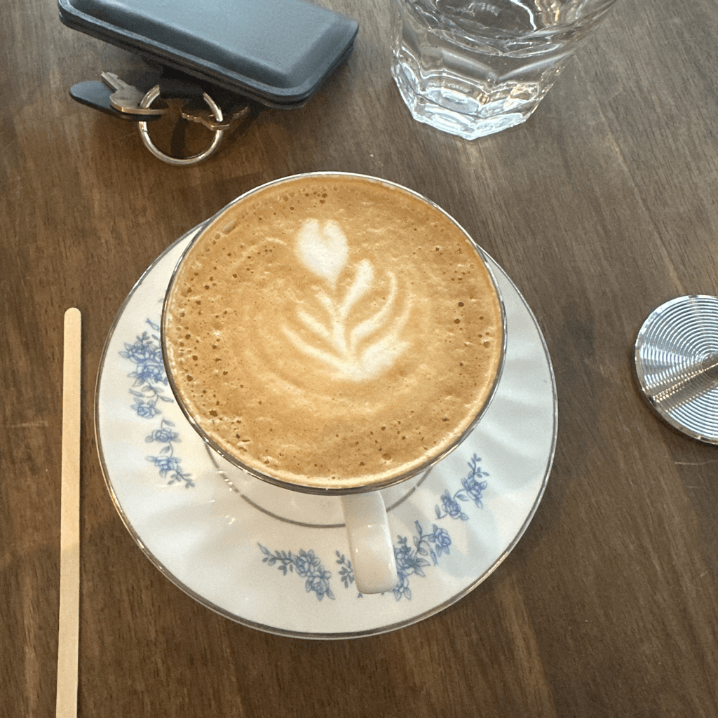 A cappuccino sits centered on a white porcelain saucer decorated with a delicate blue floral pattern and a thin silver rim, viewed from a high overhead angle on a dark brown wood-grain table.