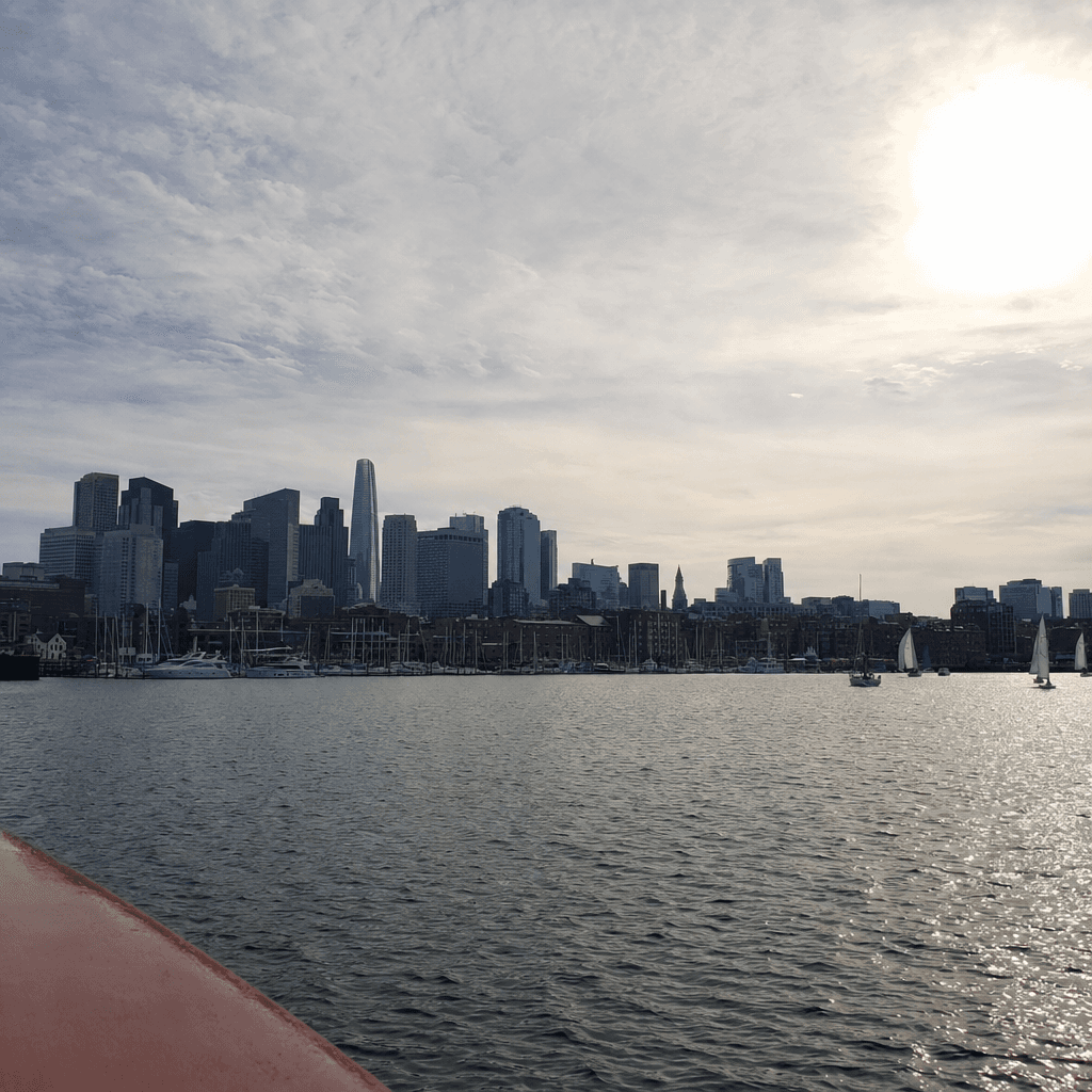 A wide waterfront view of a city skyline captured from across a broad expanse of rippling gray-blue water, likely from a boat given the sliver of reddish-brown hull visible in the lower left foreground.