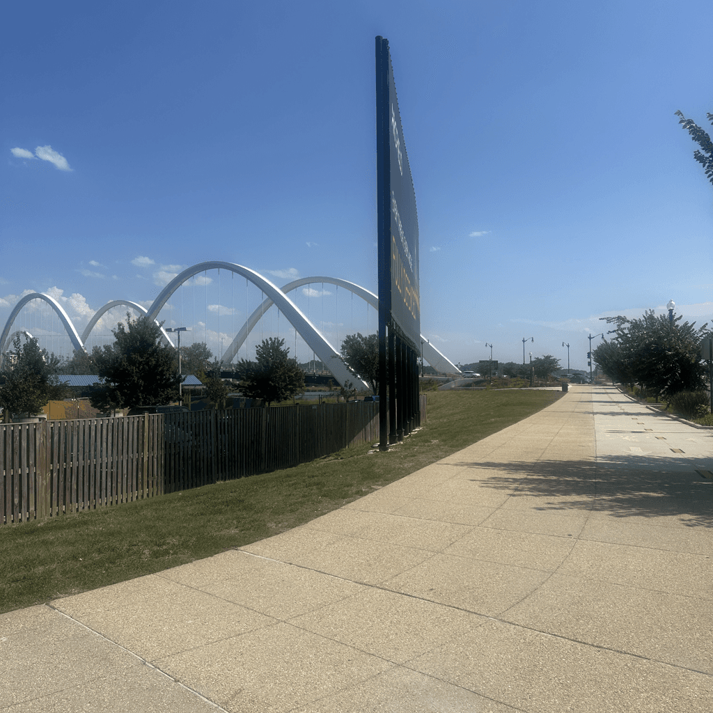 A wide paved pedestrian walkway of large concrete slabs sweeps from the lower foreground toward a vanishing point on the right, bordered on the left by a low weathered wooden picket fence and a strip of green grass.