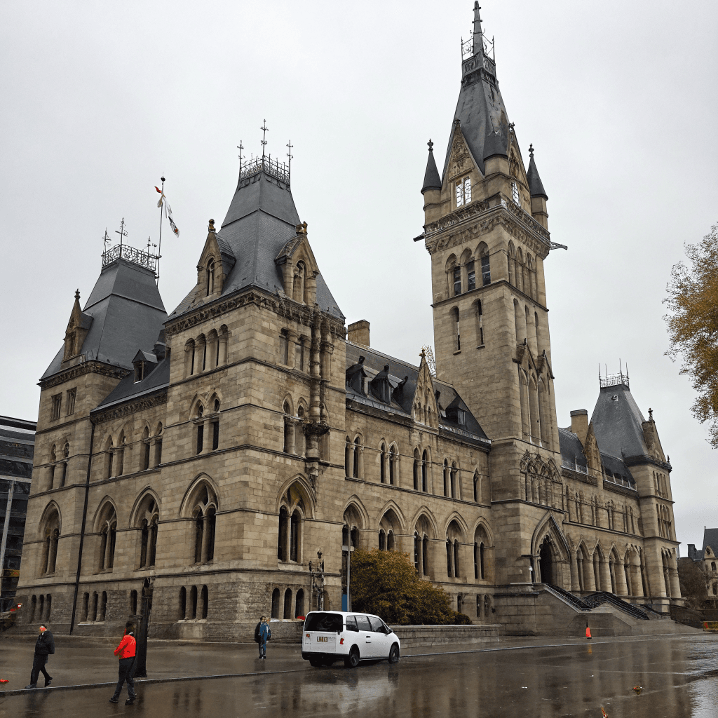 A grand Gothic-style stone building dominates the midground, its vast steep gray slate roof punctuated by dormer windows and topped by a tall central belfry tower with a clock face and pointed corner turrets, alongside slender ornate spires rising toward the right.