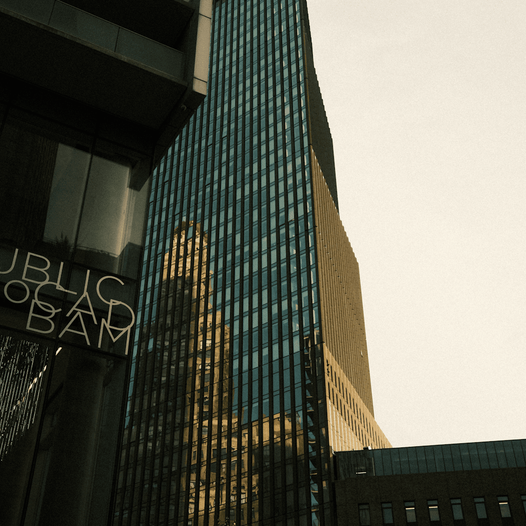 A low-angle street view looks up between modern high-rises, dominated by a tall narrow glass tower with vertical blue-green window bands and dark mullions, its façade reflecting a warm tan building beside it.