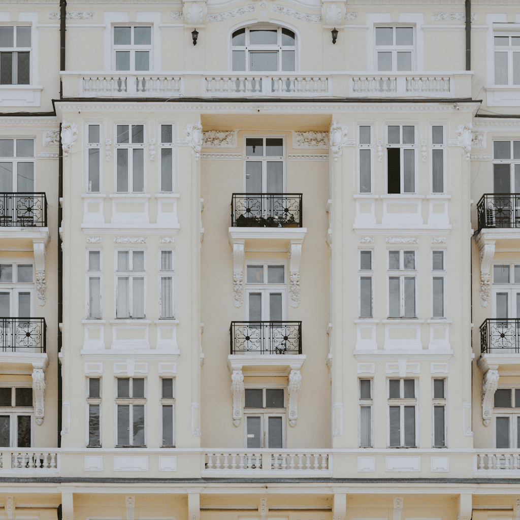 A frontal, medium-distance view of an ornate pale cream apartment façade fills the frame, with no street or sky visible, creating a tight architectural composition centered on the building’s symmetrical upper stories.