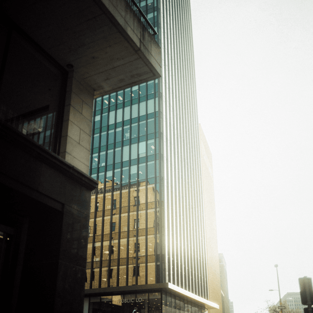 A low-angle street view looks up between modern high-rises, dominated by a tall narrow glass tower with vertical blue-green window bands and dark mullions, its façade reflecting a warm tan building beside it.