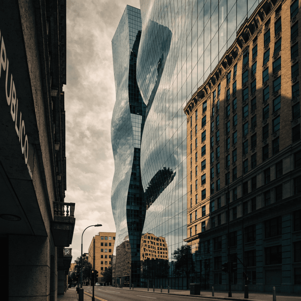 A low-angle street view looks up between modern high-rises, dominated by a tall narrow glass tower with vertical blue-green window bands and dark mullions, its façade reflecting a warm tan building beside it.