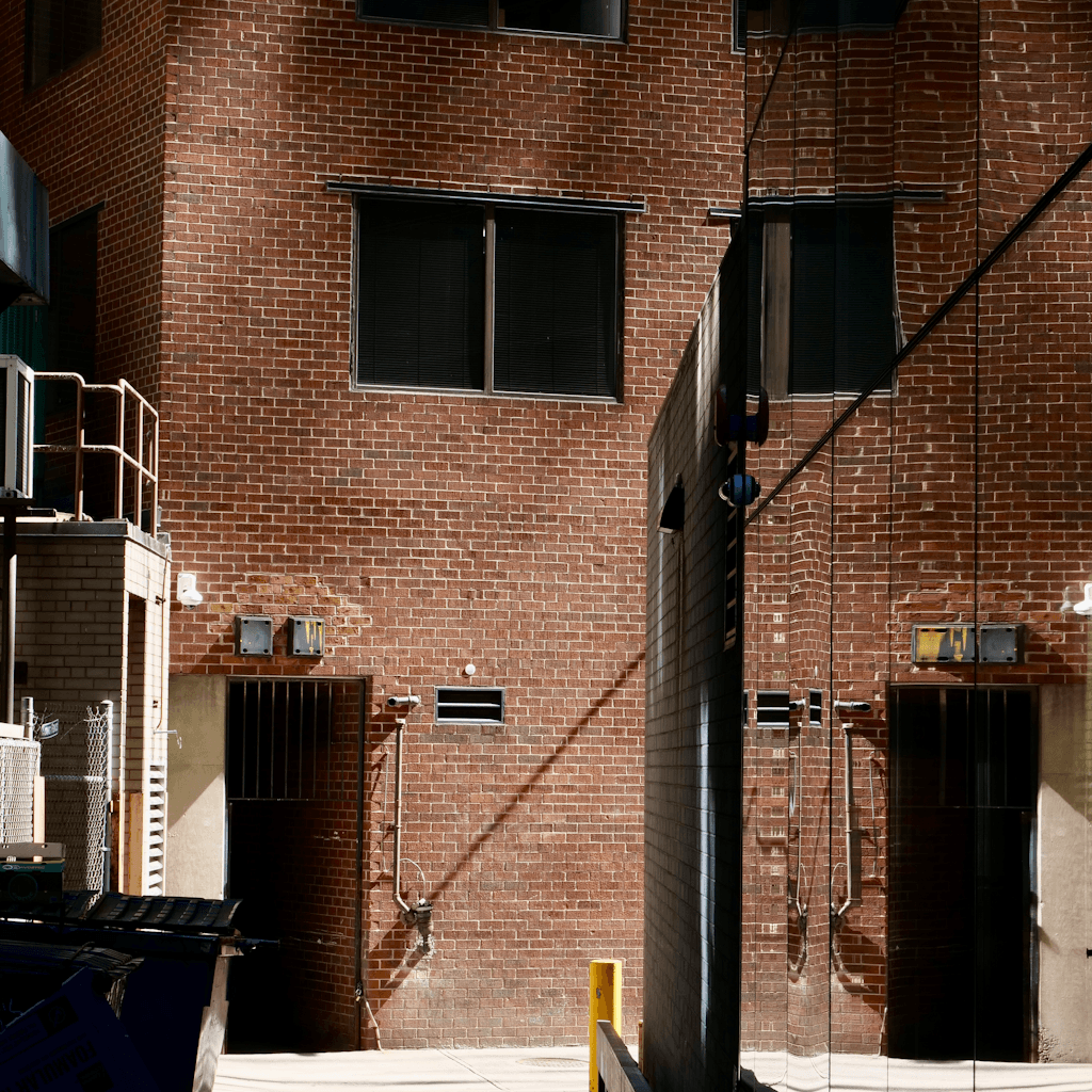 A narrow urban service alley is framed by tall red-brown brick building walls with a tight gap down the center, viewed from close range at ground level.