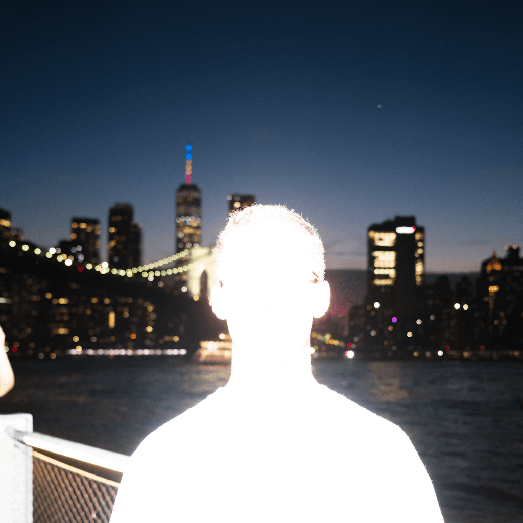 A person is silhouetted and dramatically overexposed by a bright frontal light source, appearing as a nearly white glowing form filling the lower-center foreground, with short hair backlit and the head and shoulders blocking much of the midground.