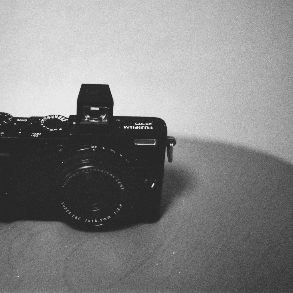 A compact black Fujifilm X70 camera sits close to the viewer on a plain, lightly textured surface, framed from a slightly elevated front-left angle so the lens and top plate dominate the lower half of the composition.