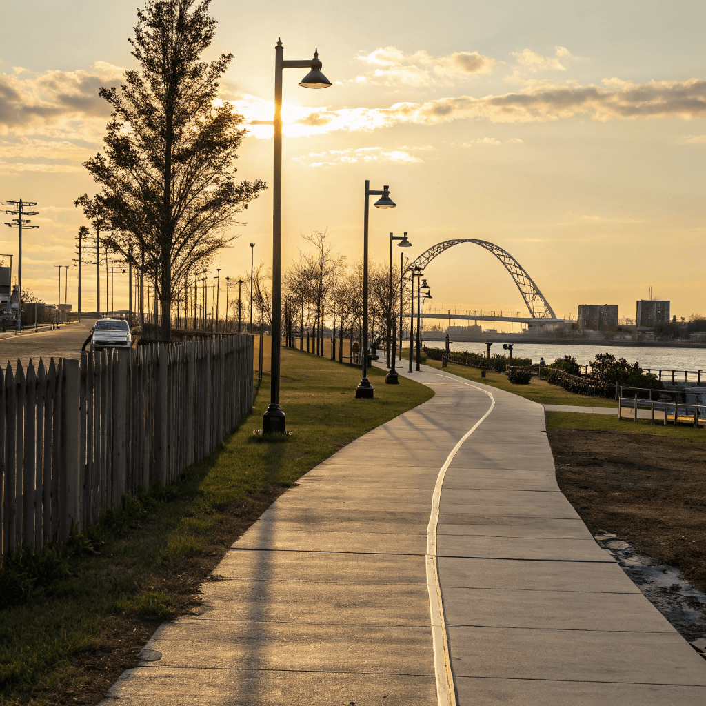 A wide paved pedestrian walkway of large concrete slabs sweeps from the lower foreground toward a vanishing point on the right, bordered on the left by a low weathered wooden picket fence and a strip of green grass.