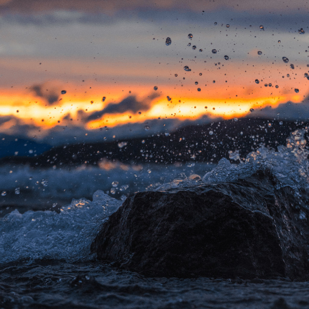 A low-angle view at the edge of dark, churning water shows a wet, rounded rock in the foreground as splashes and droplets arc upward across the frame.