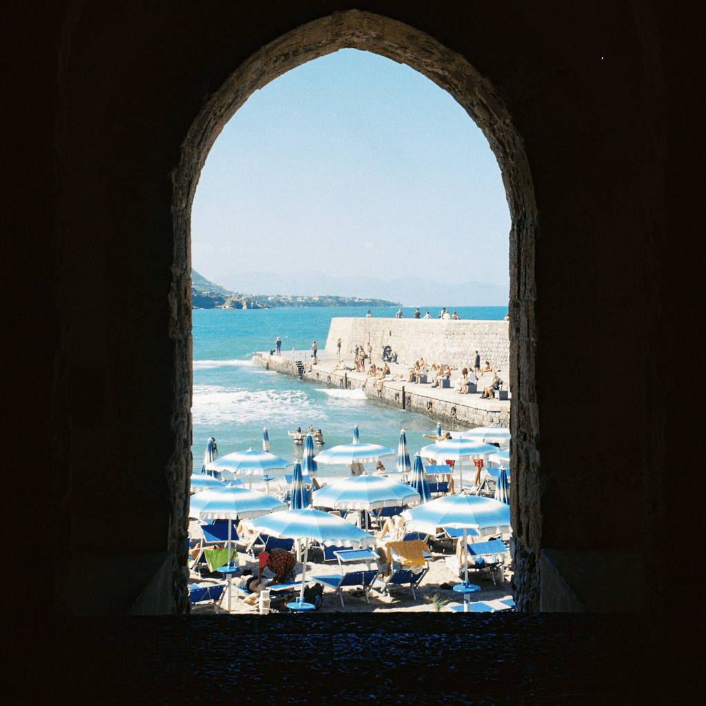 The scene unfolds outdoors, viewed through an arched stone window, framing a vibrant beach setting.
