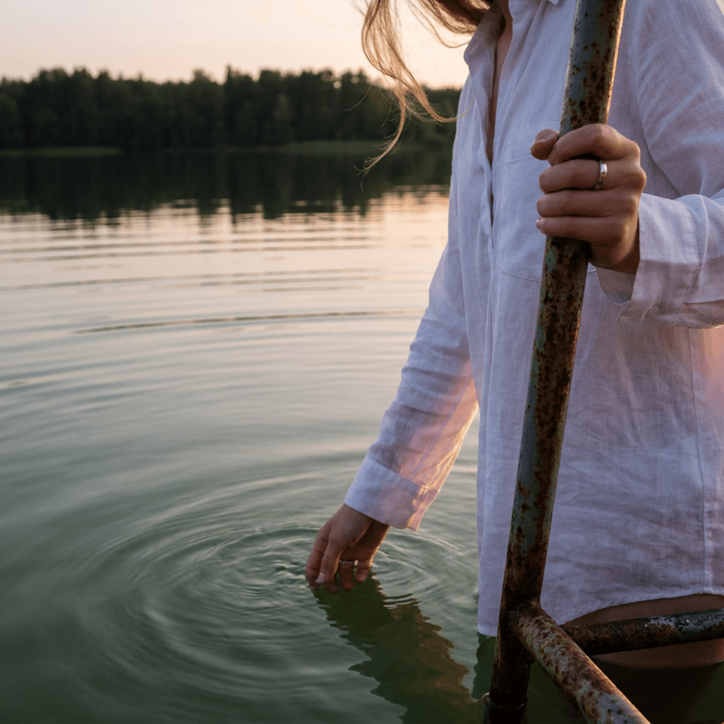 A close-up outdoor scene at the edge of calm water shows a person wading chest-deep or lower near a metal ladder, with one hand gripping a rusted vertical rail and the other hand extended into the water in the foreground.