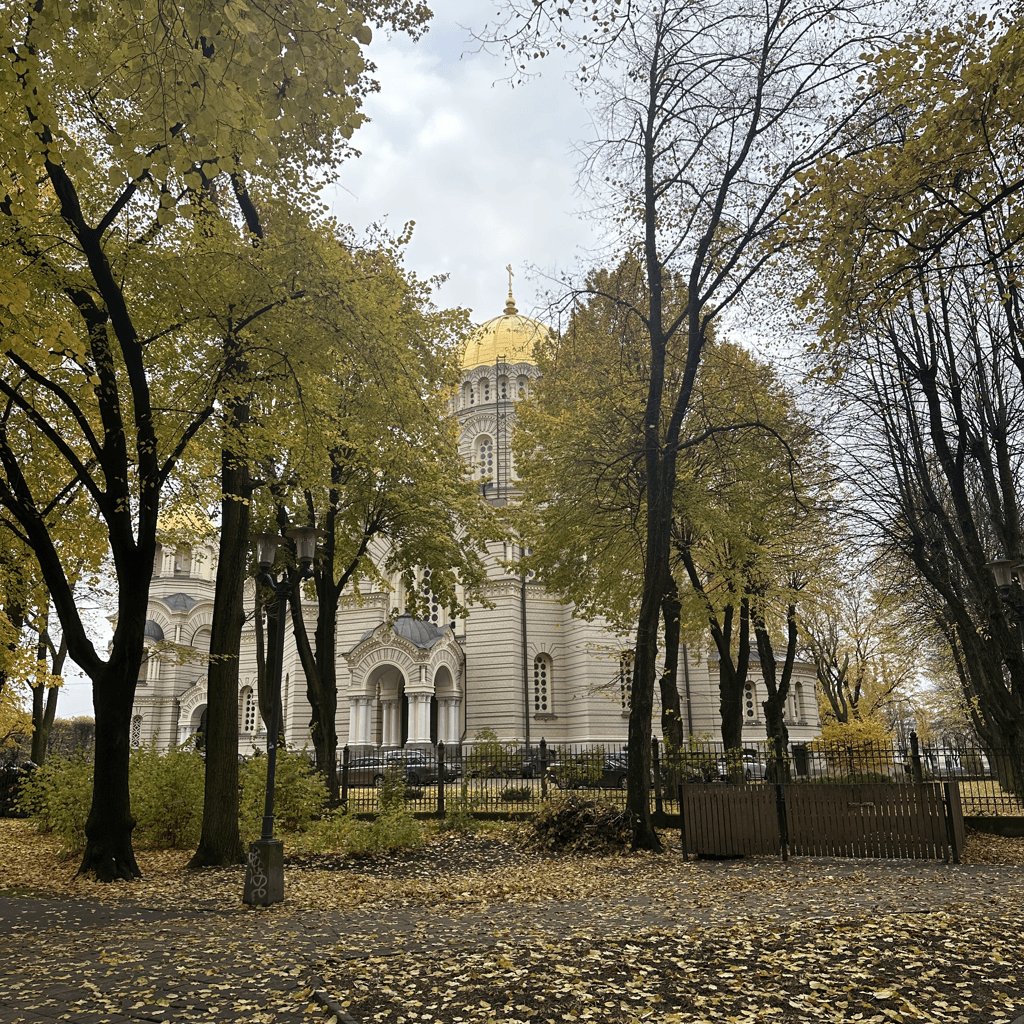 An Orthodox cathedral with a prominent golden dome topped by a cross rises in the midground, its pale stone facade banded in horizontal courses, featuring arched windows, rounded apses, and a grand arcaded entrance porch with columns.