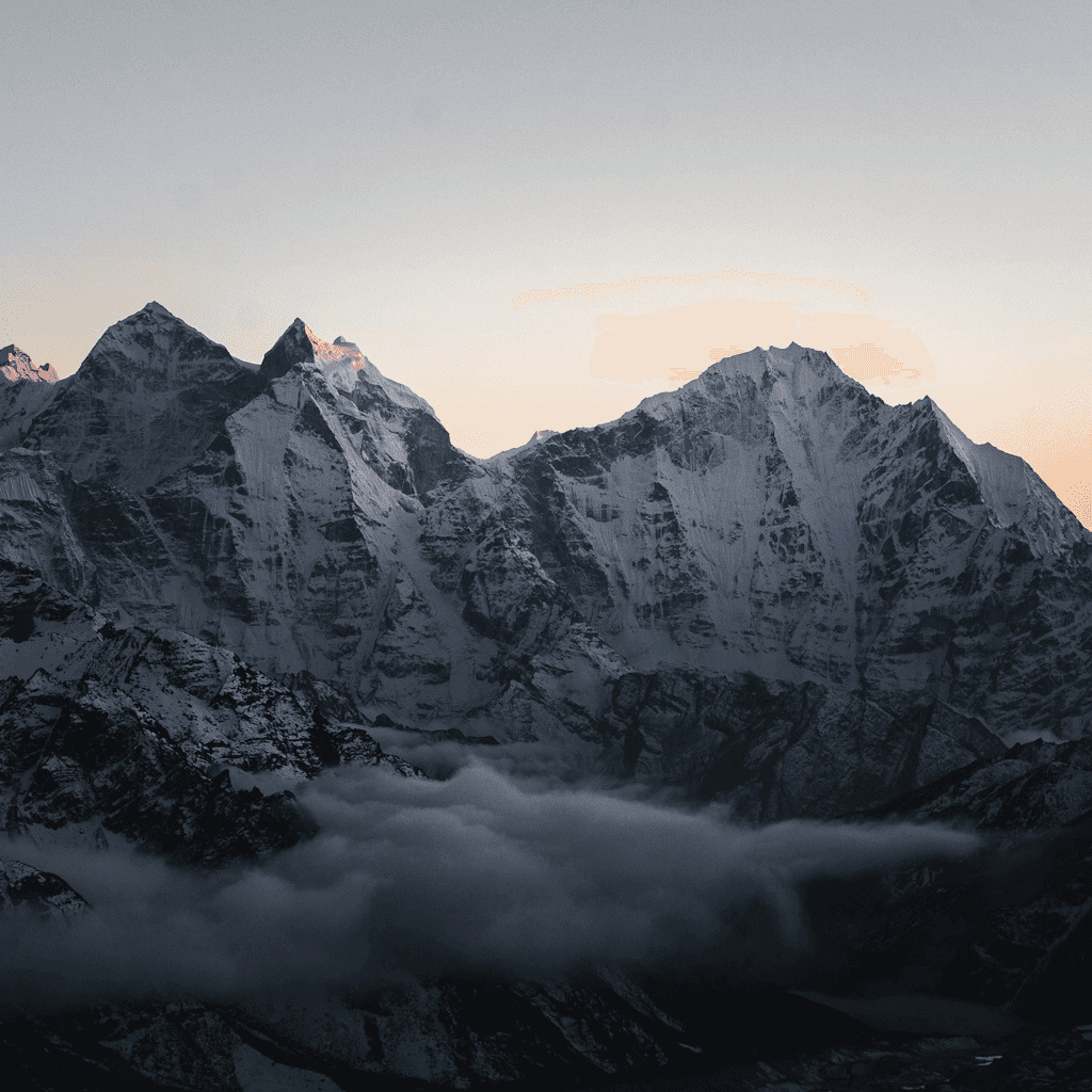 A wide, high-angle view shows a dramatic alpine mountain range filling most of the frame, with two dominant jagged snow-covered peaks rising above darker ridgelines.