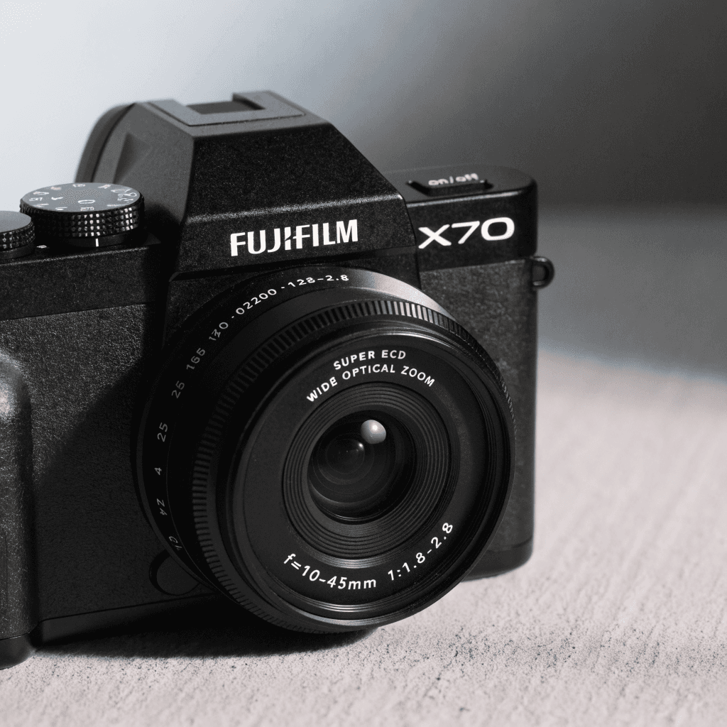 A compact black Fujifilm X70 camera sits close to the viewer on a plain, lightly textured surface, framed from a slightly elevated front-left angle so the lens and top plate dominate the lower half of the composition.