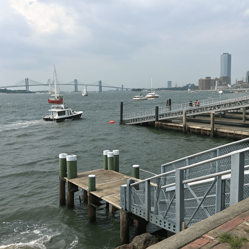 A waterfront scene captured from a pier looking across a broad expanse of gray-blue harbor toward a distant city skyline of mid-rise and high-rise buildings, some glassy and modern, softened by atmospheric haze.
