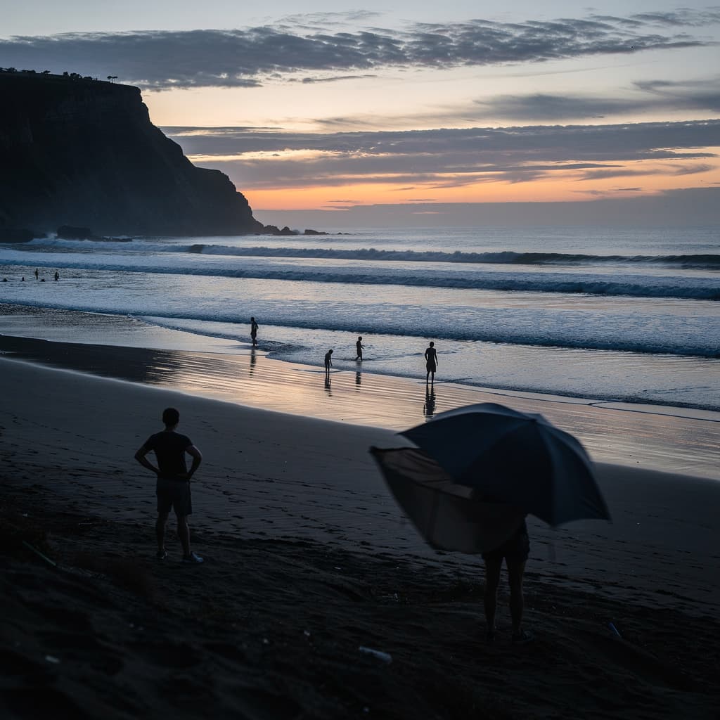 A wide seaside view at dusk shows a dark sandy beach in the foreground, with low rolling waves and a calm ocean stretching to the horizon under a pale orange band of sunset fading into blue-gray sky.