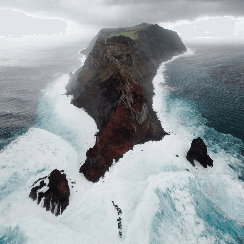 The scene presents a dramatic aerial view of an island jutting out into the ocean, framed by turbulent waves.