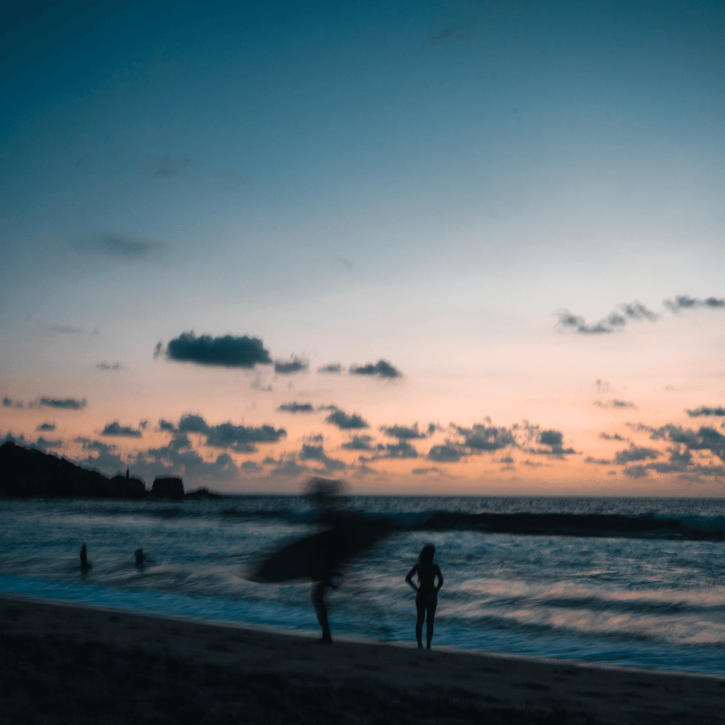 A wide seaside view at dusk shows a dark sandy beach in the foreground, with low rolling waves and a calm ocean stretching to the horizon under a pale orange band of sunset fading into blue-gray sky.