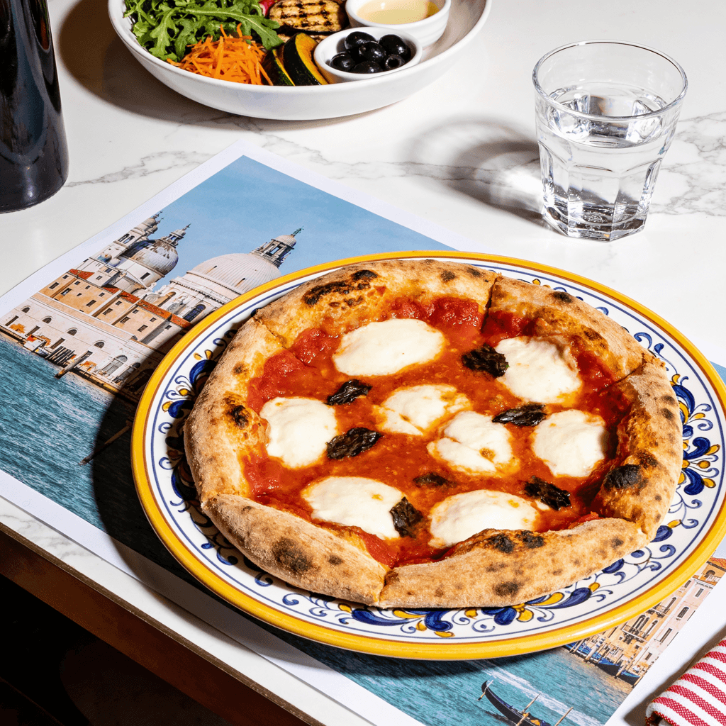 A freshly baked Neapolitan-style Margherita pizza sits centered on a decorative ceramic plate with a yellow rim and blue floral edge design, viewed from a slightly elevated angle.