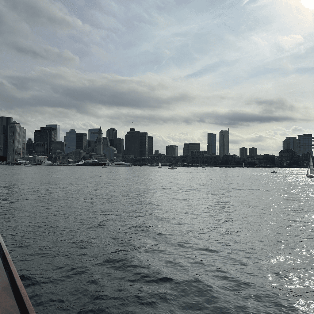 A wide waterfront view of a city skyline captured from across a broad expanse of rippling gray-blue water, likely from a boat given the sliver of reddish-brown hull visible in the lower left foreground.