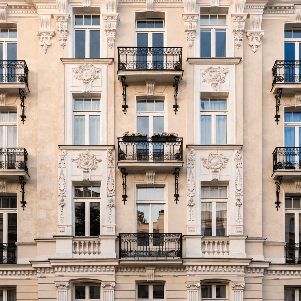 A frontal, medium-distance view of an ornate pale cream apartment façade fills the frame, with no street or sky visible, creating a tight architectural composition centered on the building’s symmetrical upper stories.