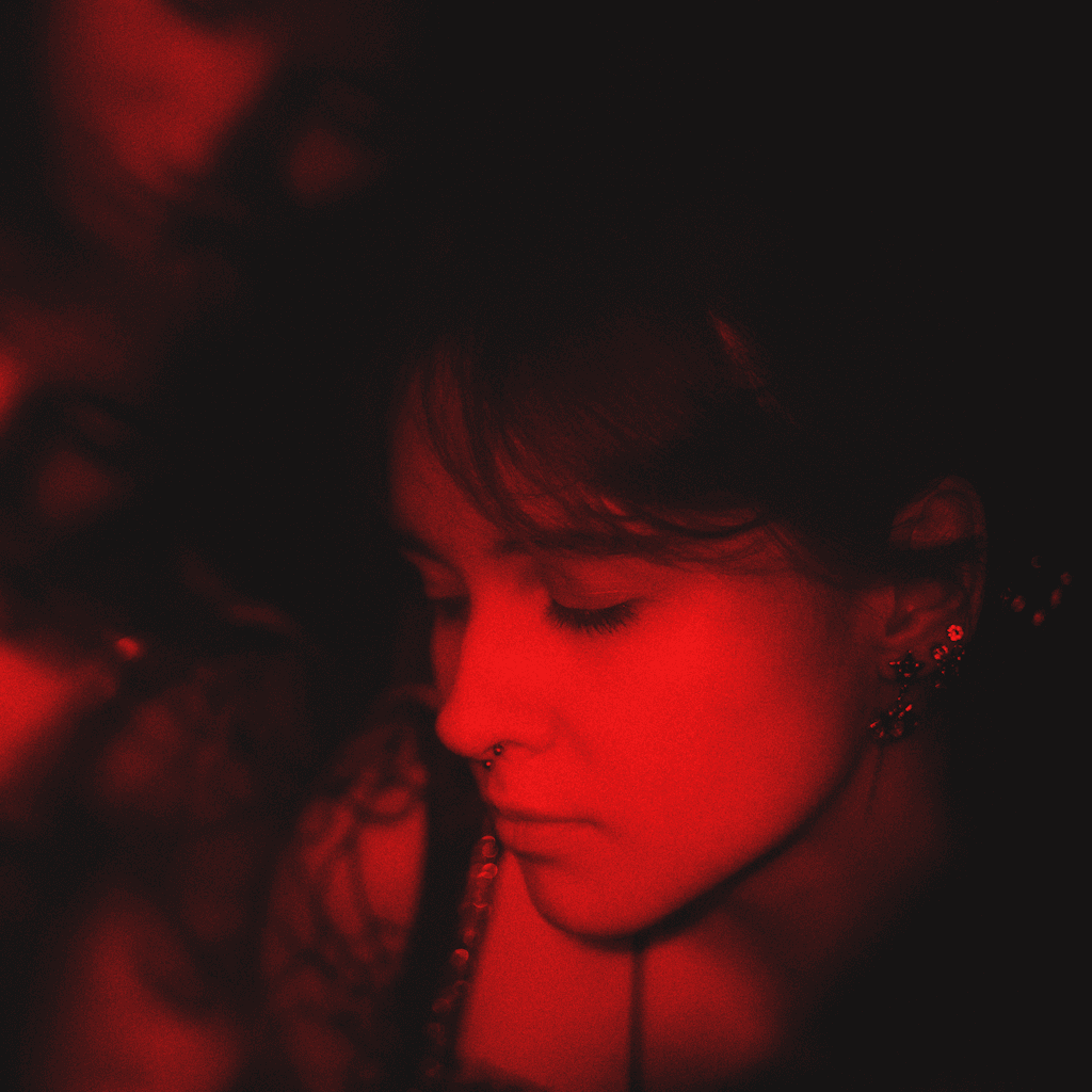 A very tight close-up shows a young woman’s face in profile turned slightly downward, filling the right half of the frame against a nearly black, indistinct background.