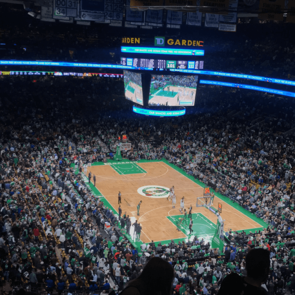 This indoor basketball arena is captured from an elevated upper-level seat, looking down at a steep angle toward the polished hardwood court in the midground, where a green-and-white team logo circle sits at center.