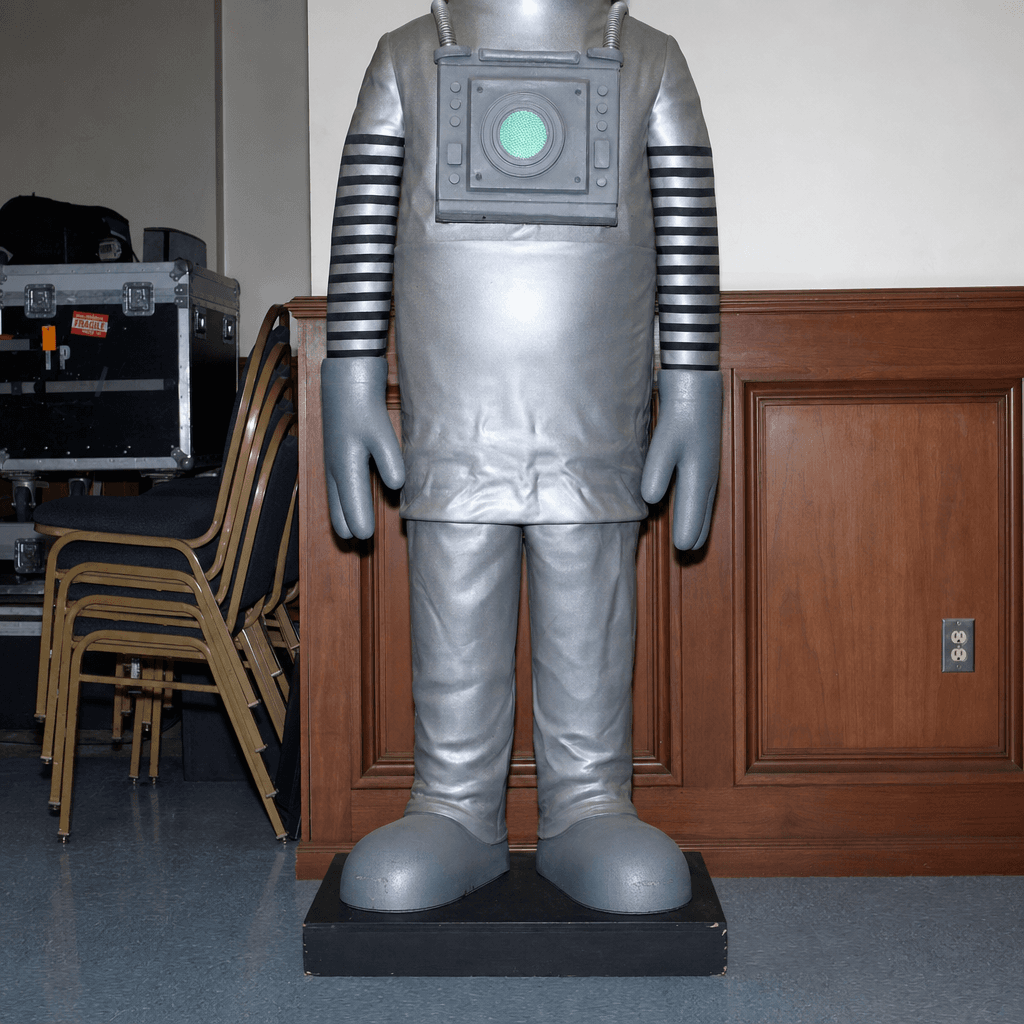A full-length novelty figure stands indoors against a mixed backdrop of wood paneling and a plain white wall, cropped at the neck and centered in the frame.