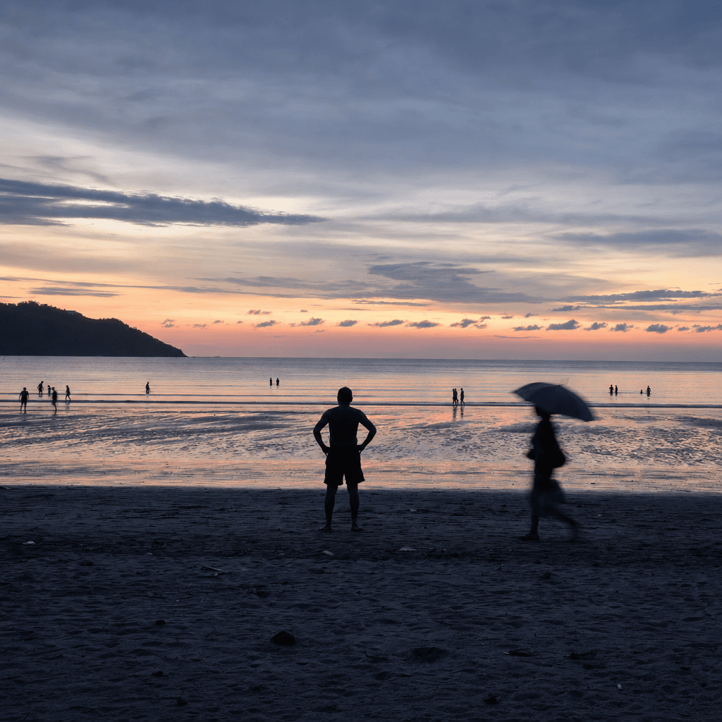 A wide seaside view at dusk shows a dark sandy beach in the foreground, with low rolling waves and a calm ocean stretching to the horizon under a pale orange band of sunset fading into blue-gray sky.