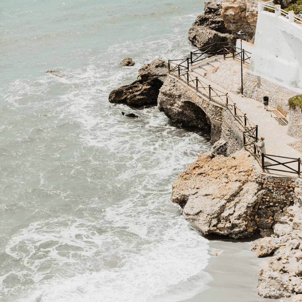 The scene is set outdoors by the sea, during a clear day with brilliant sunlight casting a shimmering glow on the turquoise water.