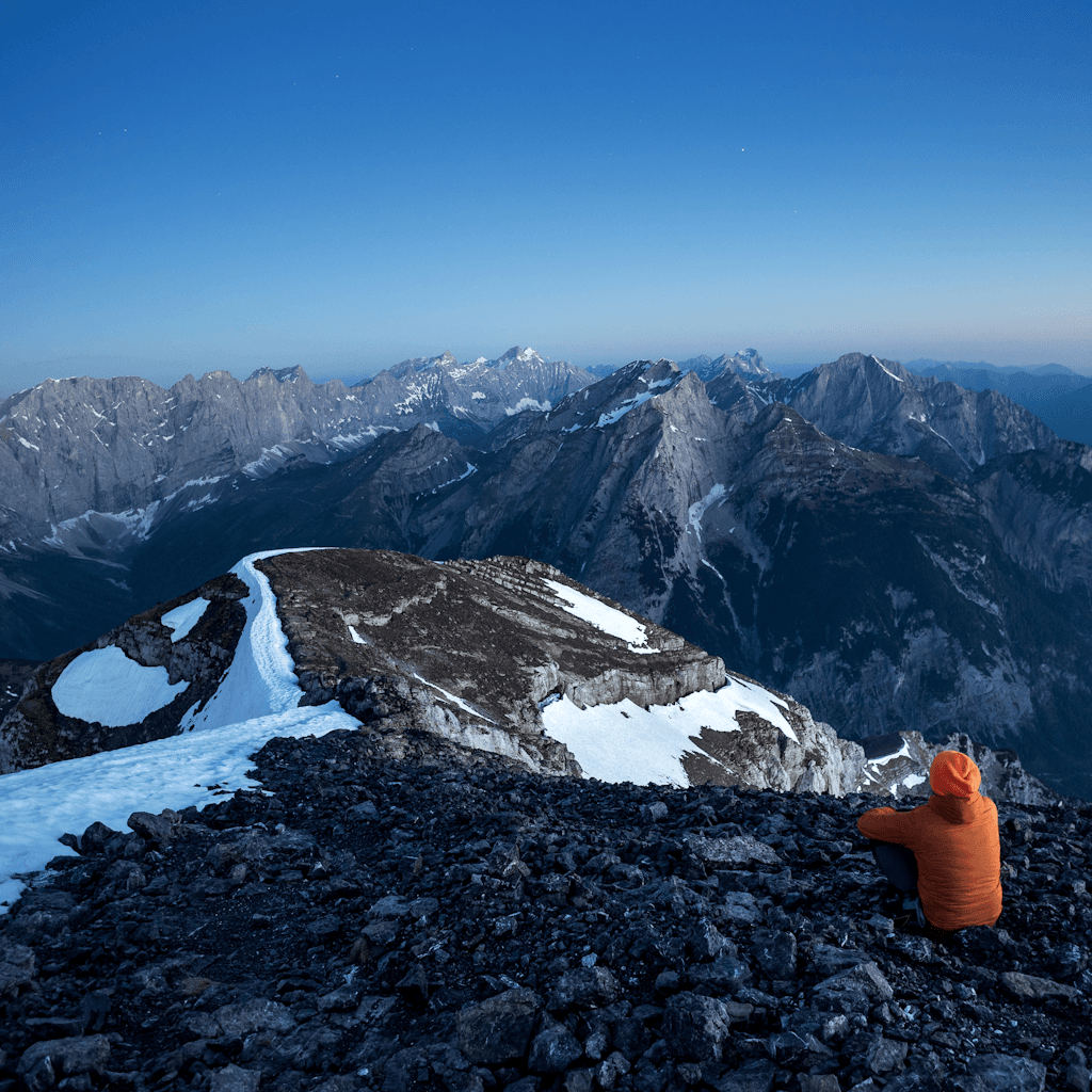A high alpine mountain scene at twilight shows a broad rocky summit in the foreground, strewn with dark scree and irregular patches of snow and ice, leading to a narrow rounded ridge.